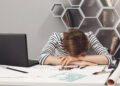 Job occupation and overwork. Close up of tired young good-looking engineer girl with dark hair in striped clothes lying on hands in office, suffering from headache after long work day