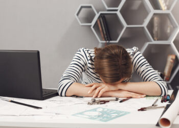 Job occupation and overwork. Close up of tired young good-looking engineer girl with dark hair in striped clothes lying on hands in office, suffering from headache after long work day