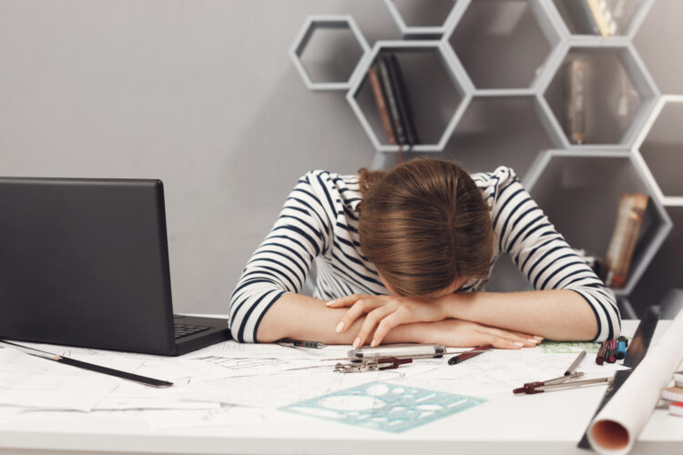 Job occupation and overwork. Close up of tired young good-looking engineer girl with dark hair in striped clothes lying on hands in office, suffering from headache after long work day