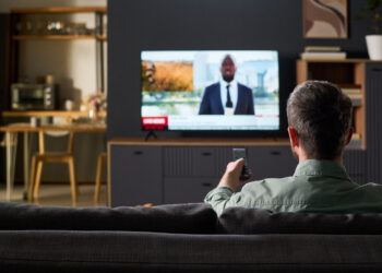 Caucasian man sitting on sofa watching television news broadcast, holding remote control and facing TV screen in modern living room interior, back view visible