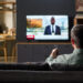 Caucasian man sitting on sofa watching television news broadcast, holding remote control and facing TV screen in modern living room interior, back view visible