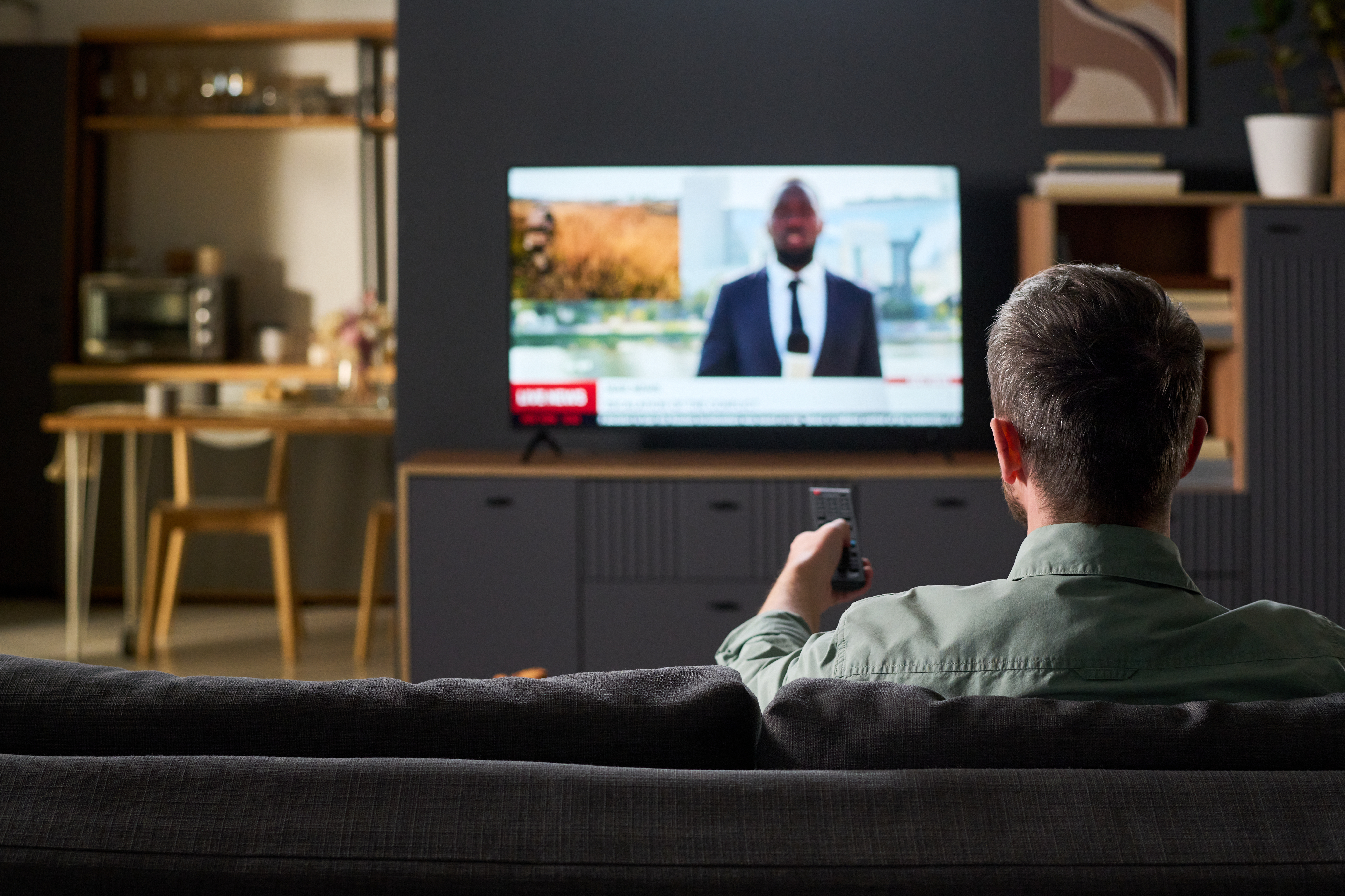 Caucasian man sitting on sofa watching television news broadcast, holding remote control and facing TV screen in modern living room interior, back view visible