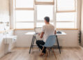 young handsome man in casual outfit sitting at table working on laptop, freelancer at home, view from back