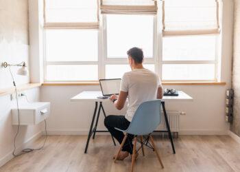 young handsome man in casual outfit sitting at table working on laptop, freelancer at home, view from back