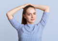 Pleasant looking young female model makes pony tail, wears striped stylish blouse, poses against blue background in studio, looks at camera with confident expression. People and beauty concept