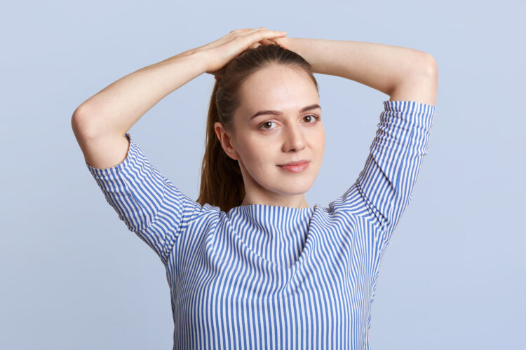 Pleasant looking young female model makes pony tail, wears striped stylish blouse, poses against blue background in studio, looks at camera with confident expression. People and beauty concept
