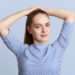 Pleasant looking young female model makes pony tail, wears striped stylish blouse, poses against blue background in studio, looks at camera with confident expression. People and beauty concept
