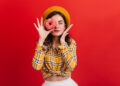 Young girl in great mood covers her eyes with strawberry donut. Woman with red lips in beret posing with closed eyes on isolated background