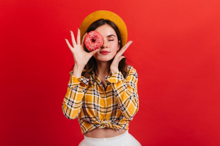 Young girl in great mood covers her eyes with strawberry donut. Woman with red lips in beret posing with closed eyes on isolated background